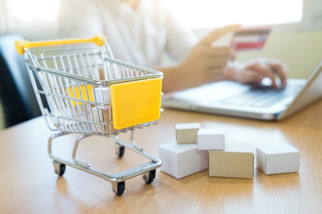 mini carro de supermercado sobre a mesa, com homem segurando um cartão de crédito e mexendo no computador no fundo da imagem